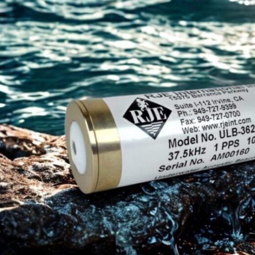 Cylindrical instrument with a white label (ULB-362B/37) resting on wet rocks beside blue sea water.