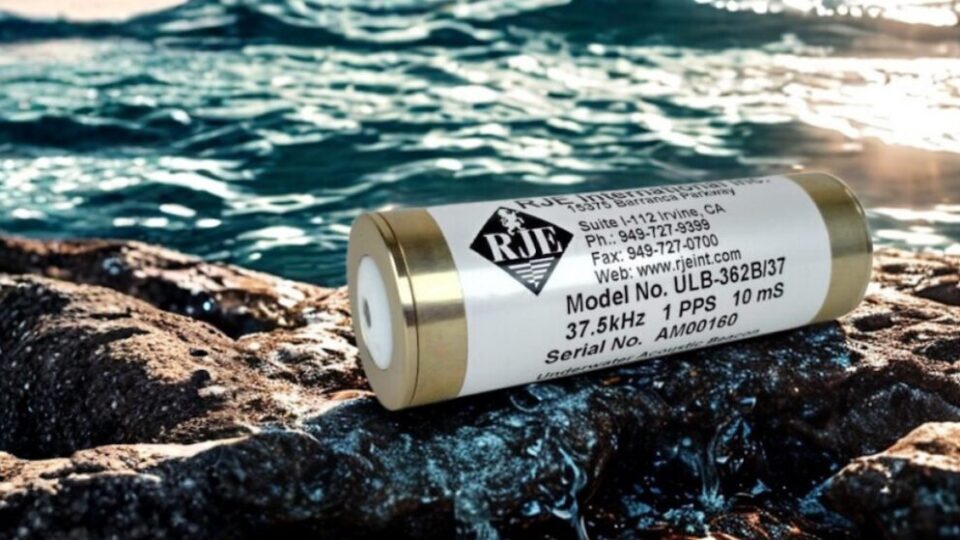 Cylindrical instrument with a white label (ULB-362B/37) resting on wet rocks beside blue sea water.