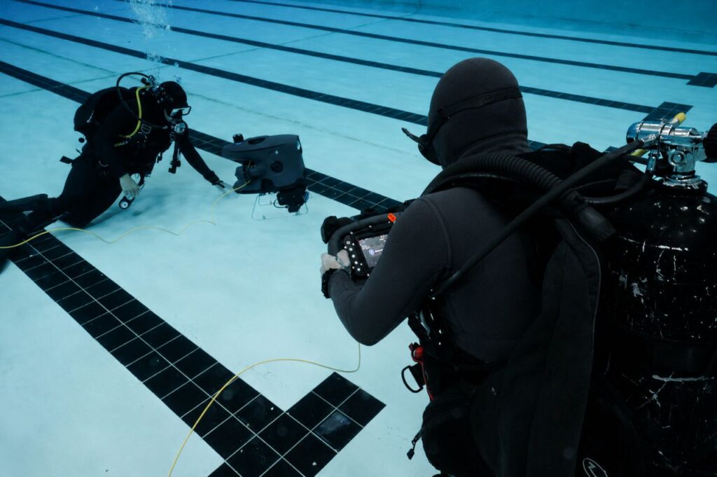 Two divers in wetsuits and dive gear operate a small remotely operated vehicle underwater in a pool.