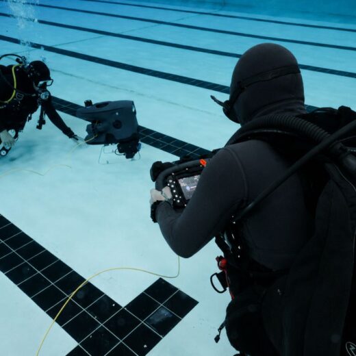 Two divers in wetsuits and dive gear operate a small remotely operated vehicle underwater in a pool.