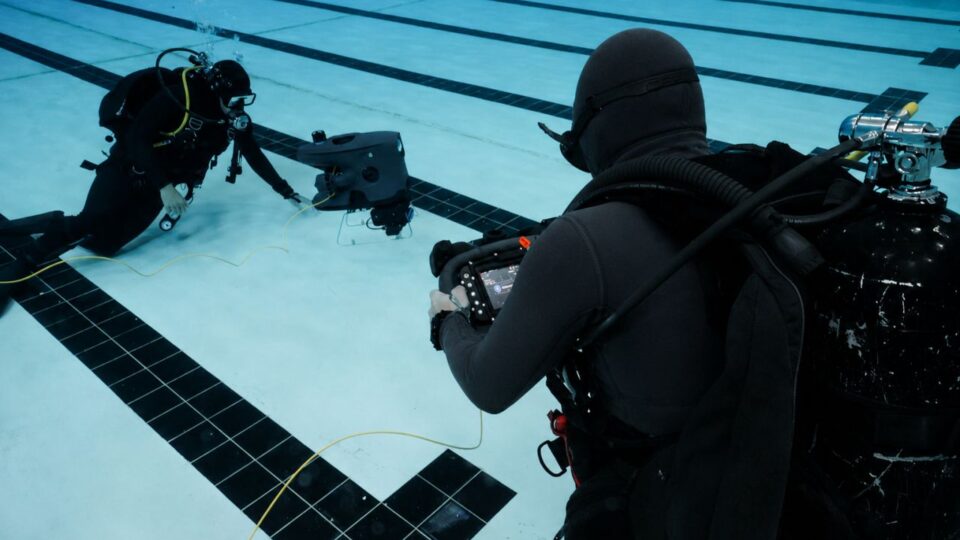 Two divers in wetsuits and dive gear operate a small remotely operated vehicle underwater in a pool.