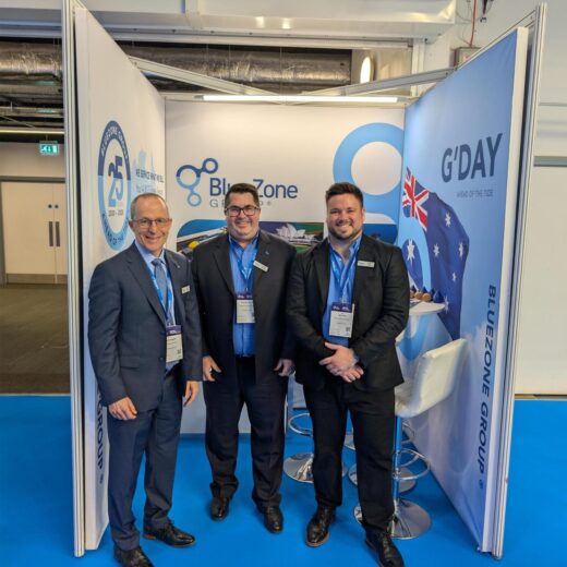 Three men in suits at a trade show booth posing for a photo in front of BlueZone and G'DAY banners, with lanyards and badges visible.