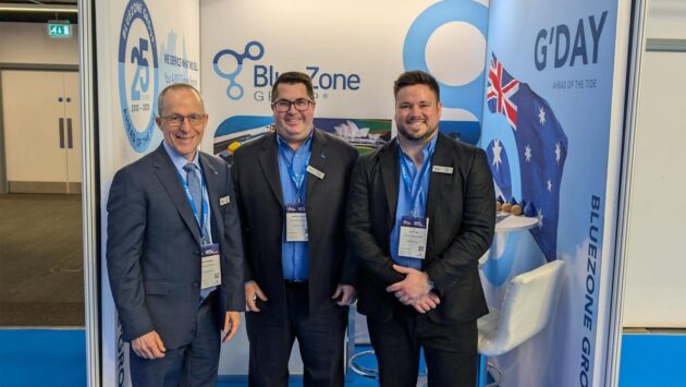 Three men in suits at a trade show booth posing for a photo in front of BlueZone and G'DAY banners, with lanyards and badges visible.