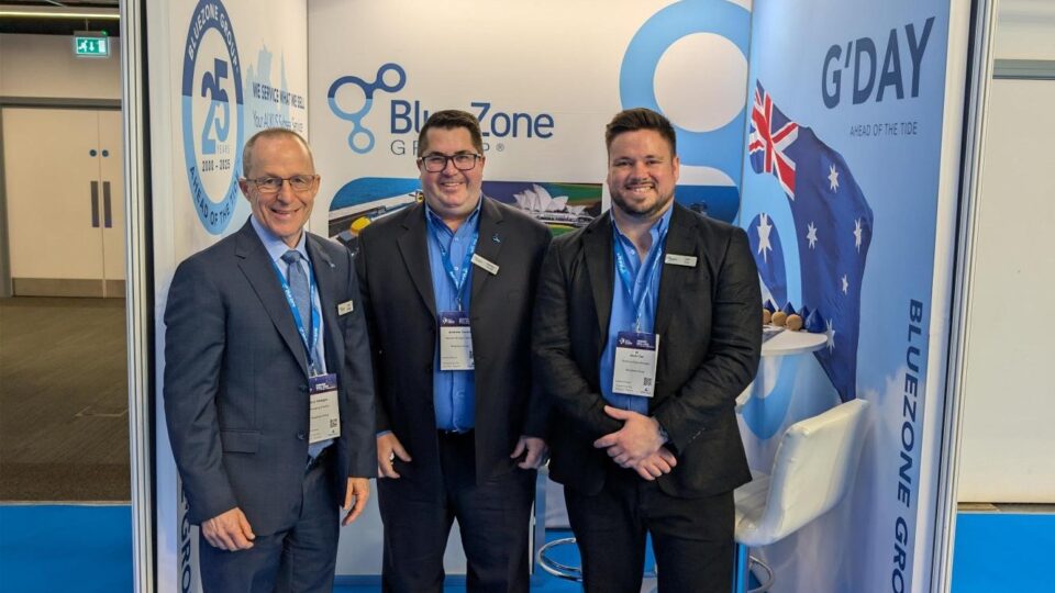 Three men in suits at a trade show booth posing for a photo in front of BlueZone and G'DAY banners, with lanyards and badges visible.