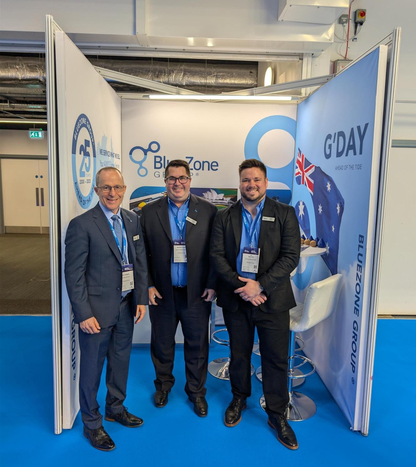 Three men in suits at a trade show booth posing for a photo in front of BlueZone and G'DAY banners, with lanyards and badges visible.