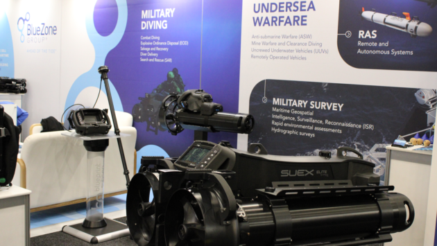Exhibit booth with blue panels titled ‘Military Diving’ and ‘Undersea Warfare’, showing a large black marine deployment device and control console on display.