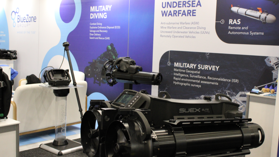 Exhibit booth with blue panels titled ‘Military Diving’ and ‘Undersea Warfare’, showing a large black marine deployment device and control console on display.
