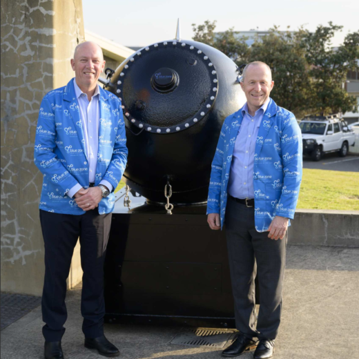 Two men in matching blue jackets stand beside a large black industrial vessel with a riveted circular front outdoors.