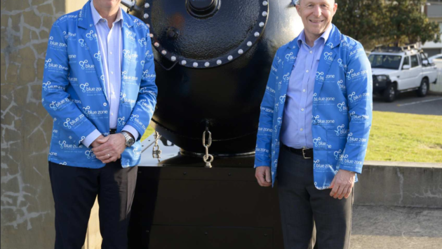 Two men in matching blue jackets stand beside a large black industrial vessel with a riveted circular front outdoors.
