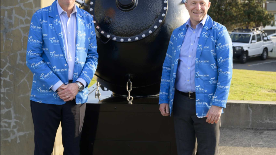 Two men in matching blue jackets stand beside a large black industrial vessel with a riveted circular front outdoors.