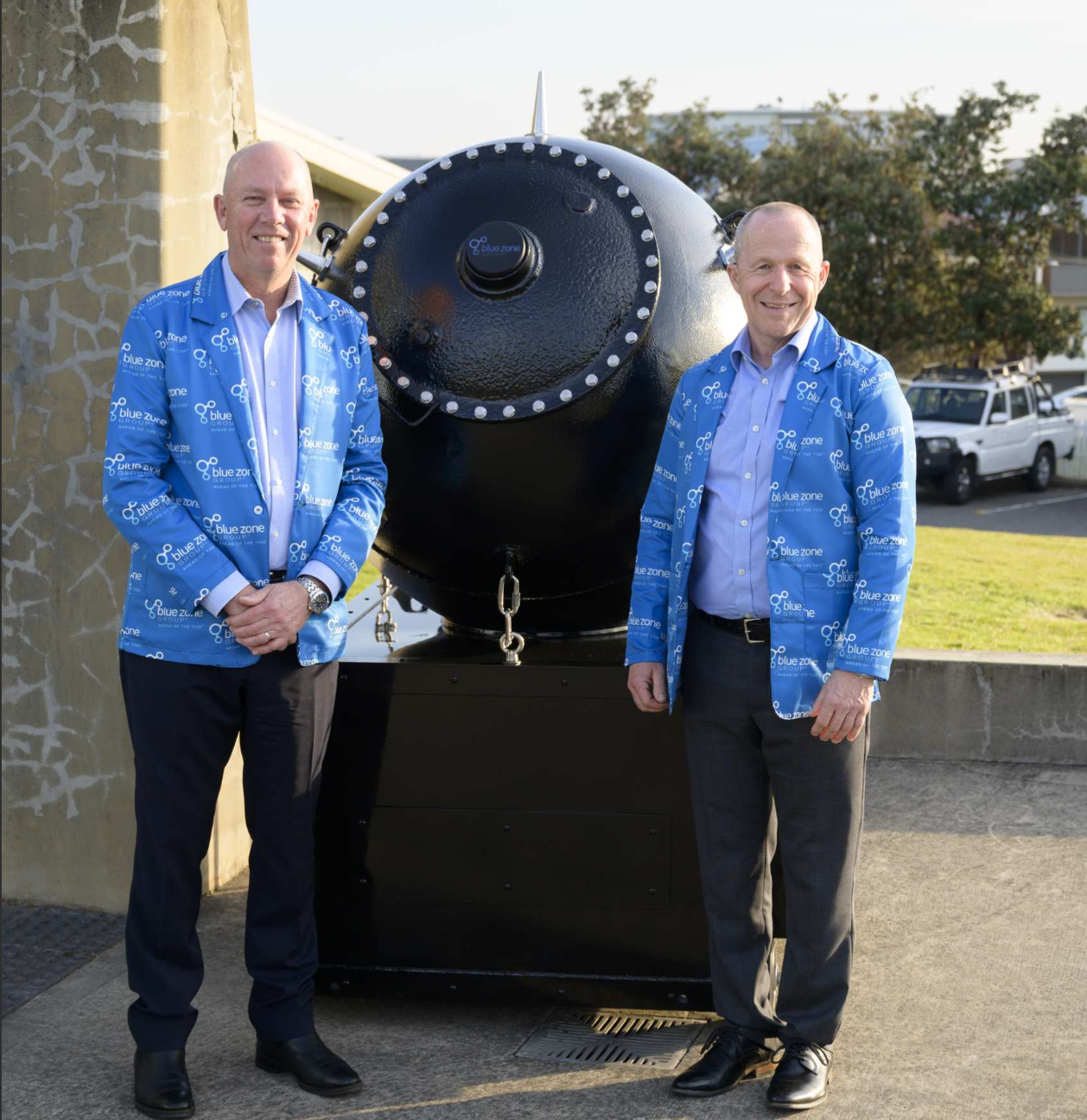 Screenshot 2026-07-06 152740 - BlueZone Group Two men in matching blue jackets stand beside a large black industrial vessel with a riveted circular front outdoors.