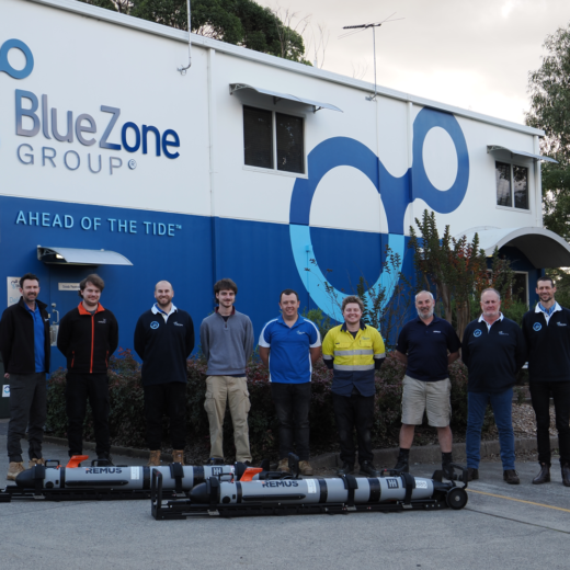 Ten employees stand in a line outside the BlueZone Group building, with a blue wave mural and the slogan 'Ahead of the Tide' visible, and several large cylindrical devices on the ground in front of them.