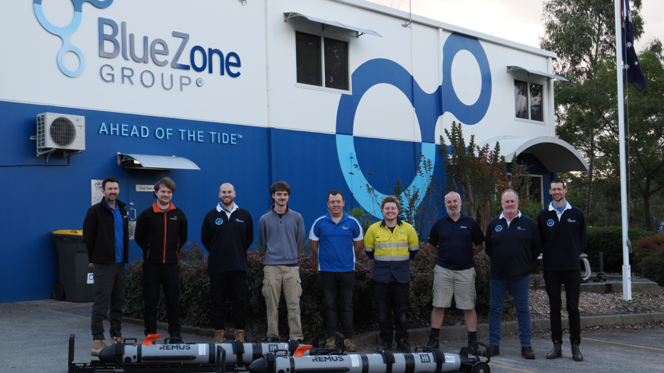 Ten employees stand in a line outside the BlueZone Group building, with a blue wave mural and the slogan 'Ahead of the Tide' visible, and several large cylindrical devices on the ground in front of them.
