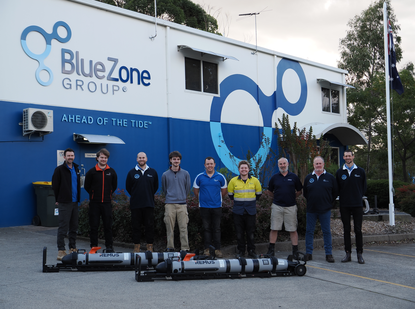 Ten employees stand in a line outside the BlueZone Group building, with a blue wave mural and the slogan 'Ahead of the Tide' visible, and several large cylindrical devices on the ground in front of them.