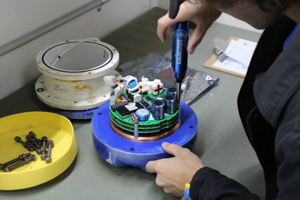 Technician using a screwdriver to assemble an electronic circuit board mounted on a blue circular base, with a yellow parts tray nearby.