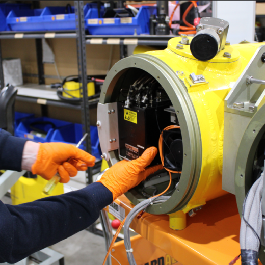 Technician wearing orange gloves works on the inside of a bright yellow industrial machine, wiring and components exposed in a workshop.