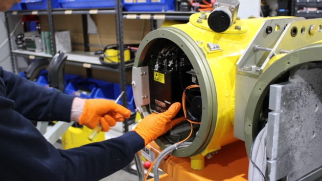 Technician wearing orange gloves works on the inside of a bright yellow industrial machine, wiring and components exposed in a workshop.