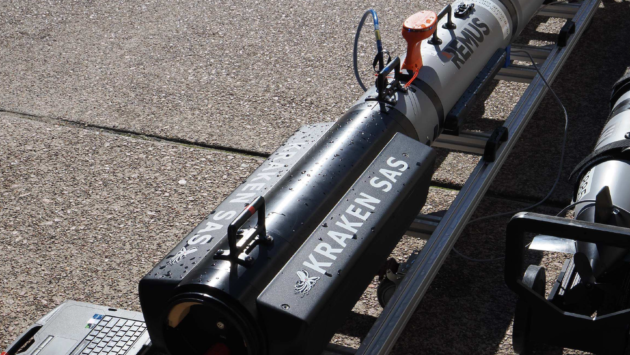 Remote-operated underwater vehicle (REMUS) and equipment on a rack with a laptop on a paved surface, connected by cables for testing/training.
