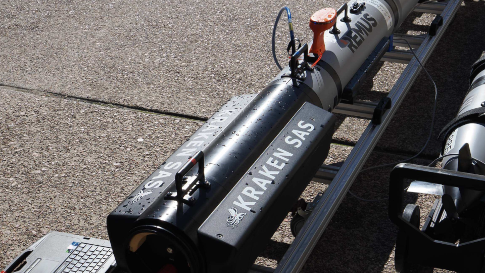 Remote-operated underwater vehicle (REMUS) and equipment on a rack with a laptop on a paved surface, connected by cables for testing/training.