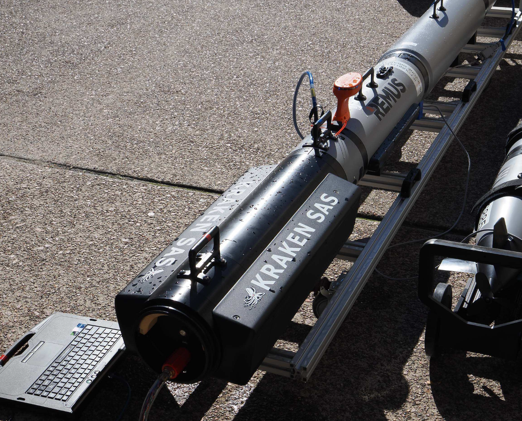 Remote-operated underwater vehicle (REMUS) and equipment on a rack with a laptop on a paved surface, connected by cables for testing/training.
