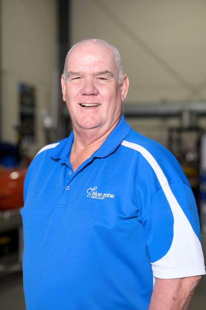 Smiling older man in a blue polo shirt with white stripes, standing in a workshop