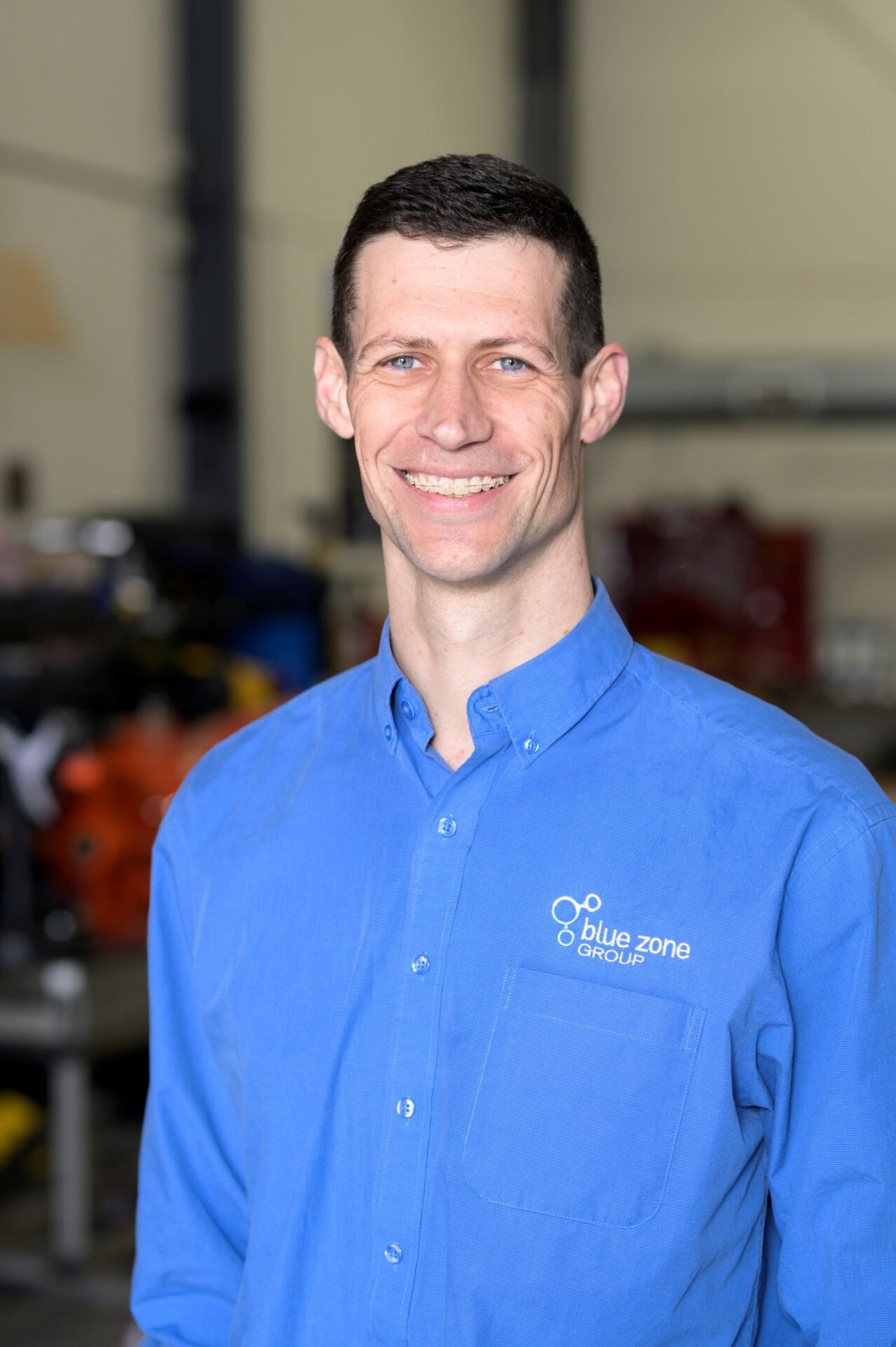 Smiling man in a blue shirt with Blue Zone Group logo, standing in an industrial workshop.