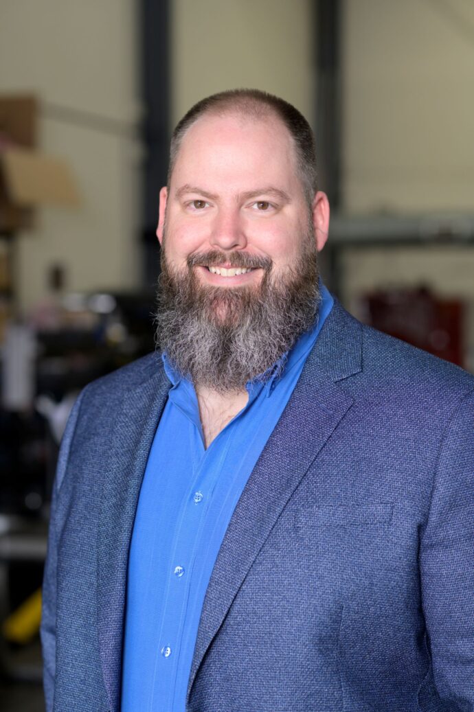 Portrait of a smiling man with a full beard wearing a blue shirt and textured blazer in a neutral indoor setting.