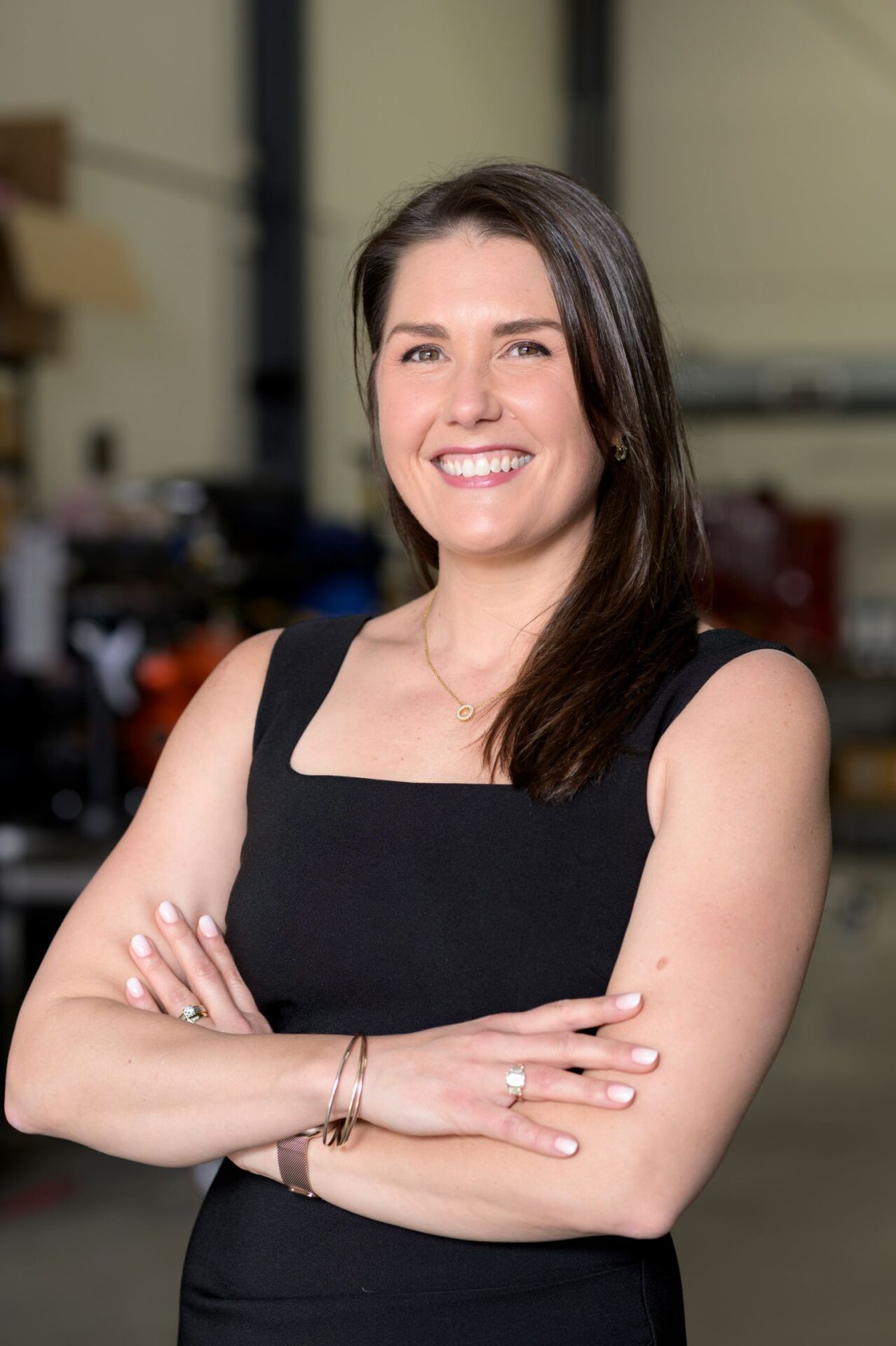 Smiling woman with dark hair in a black sleeveless dress stands with arms crossed in a workshop setting.