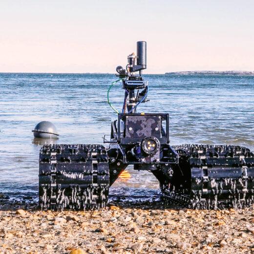 A rugged robotic vehicle with tank-like tracks sits on a rocky shoreline facing the sea, with complex equipment mounted on its frame.
