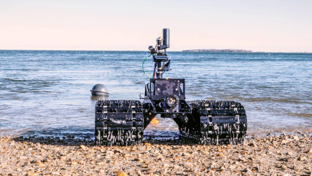 A rugged robotic vehicle with tank-like tracks sits on a rocky shoreline facing the sea, with complex equipment mounted on its frame.