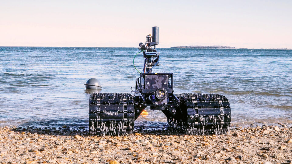 A rugged robotic vehicle with tank-like tracks sits on a rocky shoreline facing the sea, with complex equipment mounted on its frame.