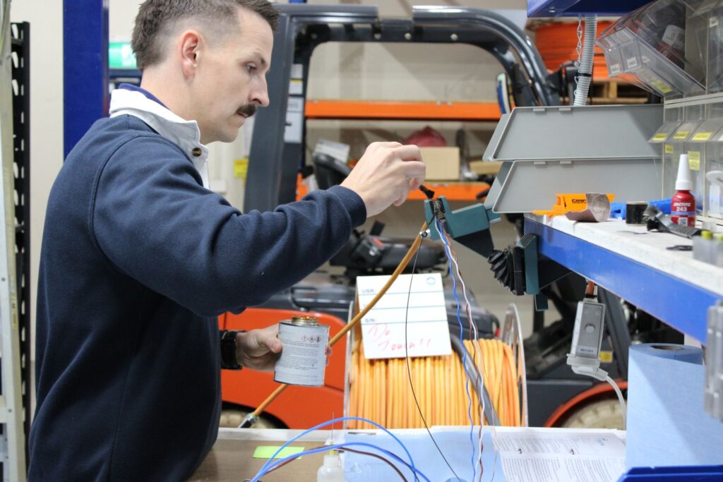 Technician in a blue sweater wires up a panel with colorful cables in a workshop, holding a tool and a can of soldering flux nearby.