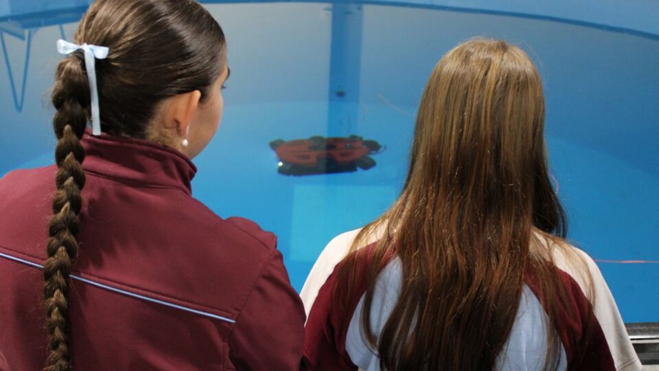 Two girls with long hair stand at an aquarium viewing panel, watching a red object float in a blue tank water.