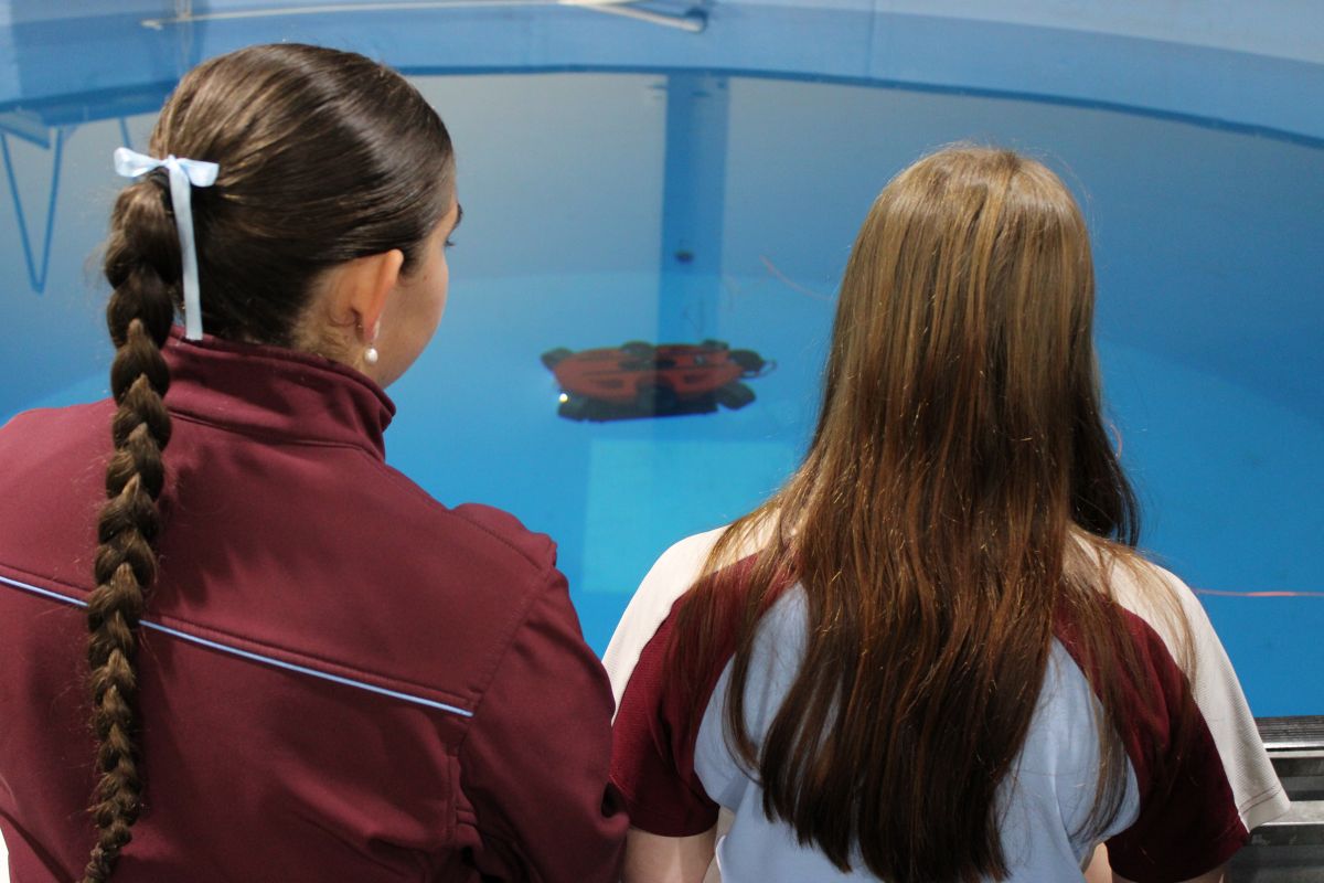 Two girls with long hair stand at an aquarium viewing panel, watching a red object float in a blue tank water.