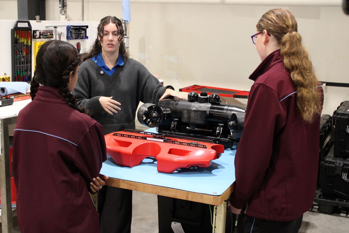 Instructor explaining a large black mechanical device on a worktable to two students in a workshop setting.