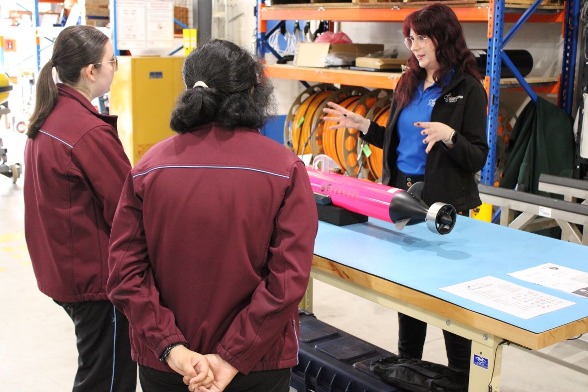 Instructor explains a pink torpedo-shaped device to two visitors in a workshop setting on a blue worktable.