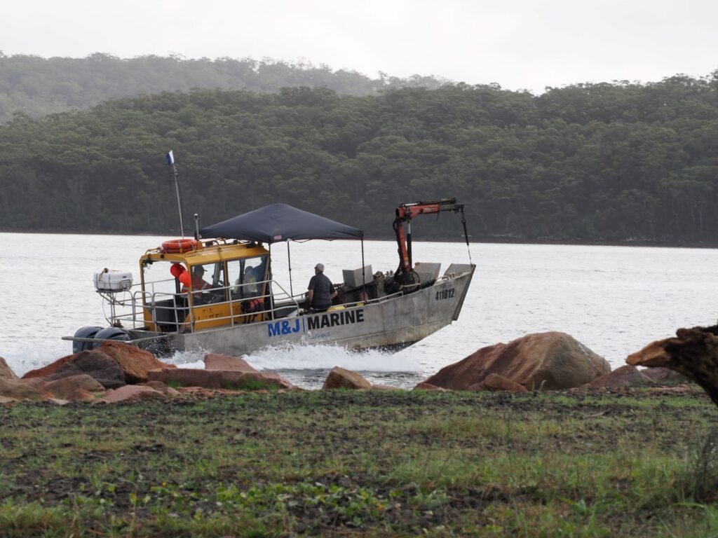 Small work boat with a yellow cabin and a crane, titled M&J MARINE, sailing near rocky shore with crew onboard under a canopy