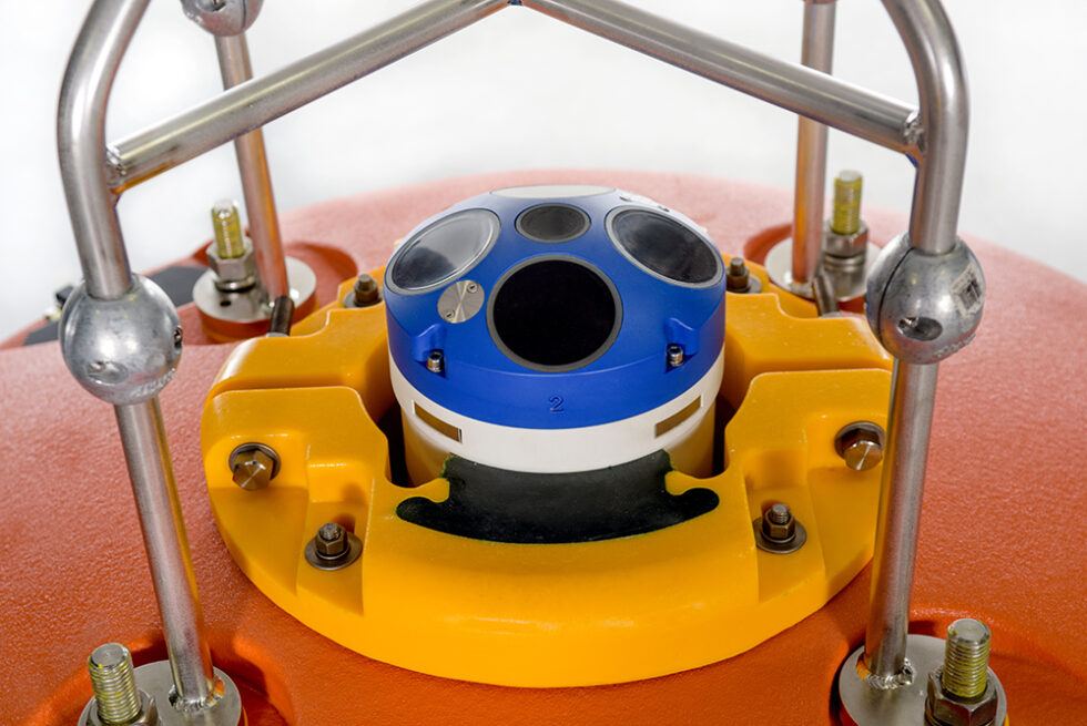 Elliptical-ADCP-Buoys-with-Teledyne-Sentinel-close-up-980x655 - BlueZone Group Blue circular sensor housing mounted in a yellow robotic base, surrounded by metal framing bars (industrial sensor setup).