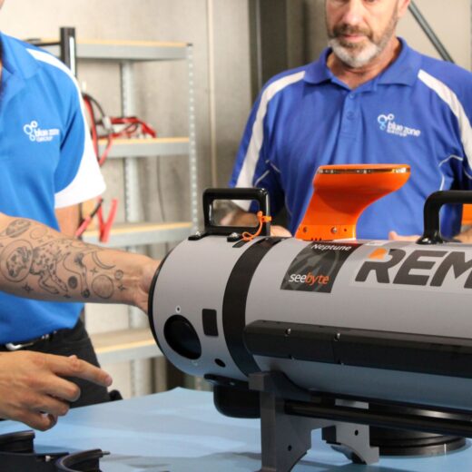 Two technicians in blue shirts position a gray REMUS underwater drone with an orange top docking sensor in a workshop.