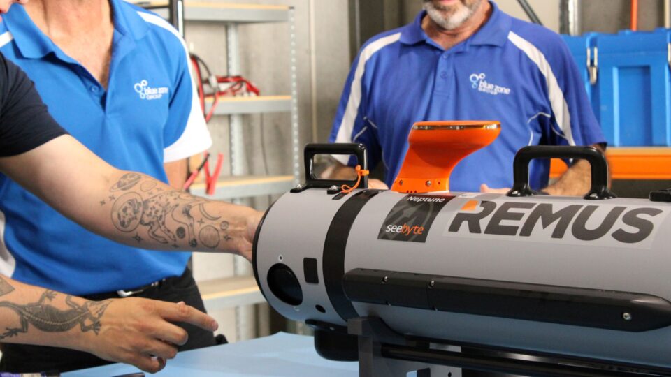 Two technicians in blue shirts position a gray REMUS underwater drone with an orange top docking sensor in a workshop.