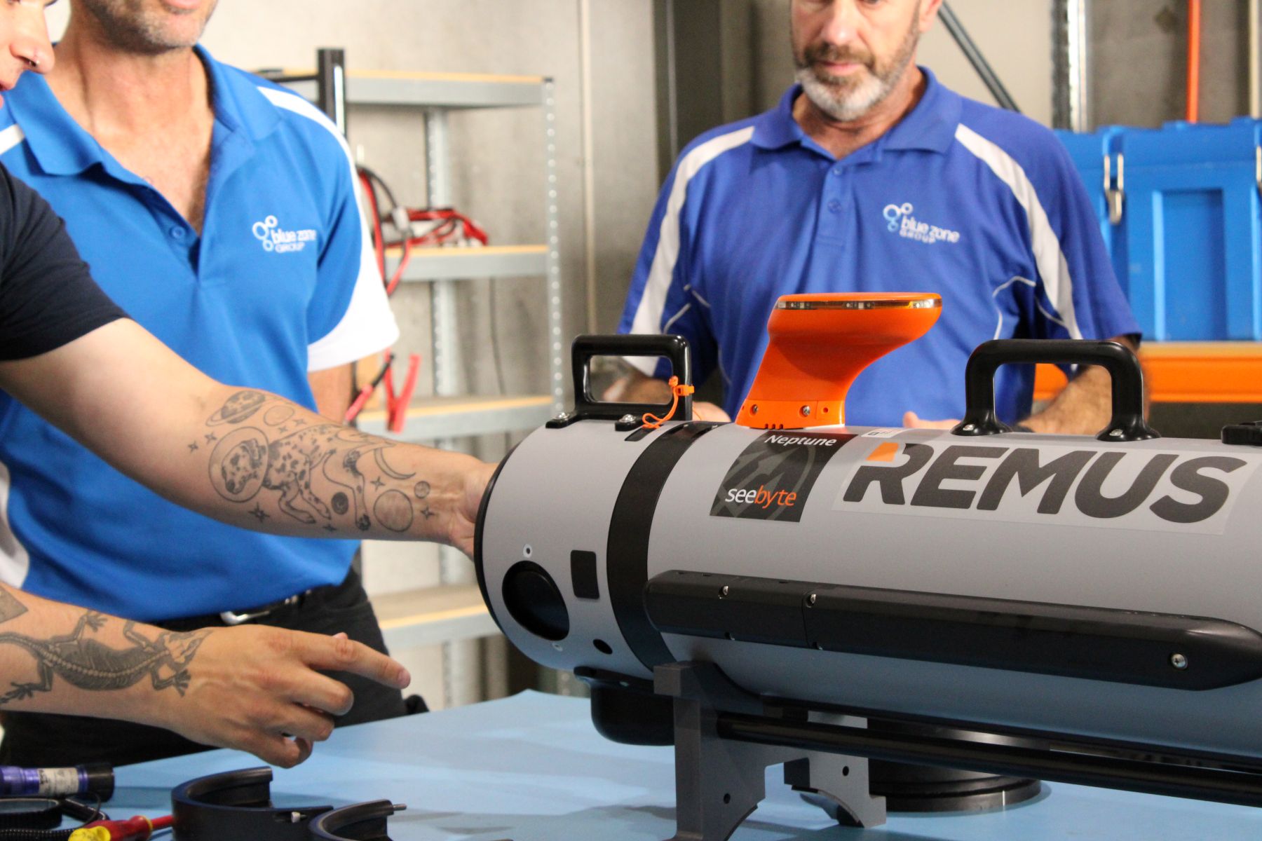 Two technicians in blue shirts position a gray REMUS underwater drone with an orange top docking sensor in a workshop.