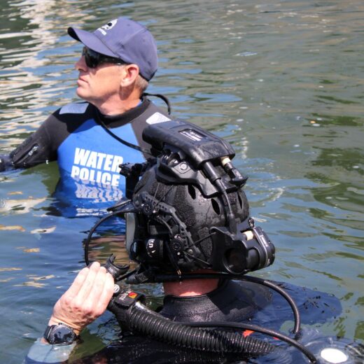 Two divers in open water: one wearing a blue 'Water Police' vest and cap, the other in a heavy diving helmet and breathing gear.