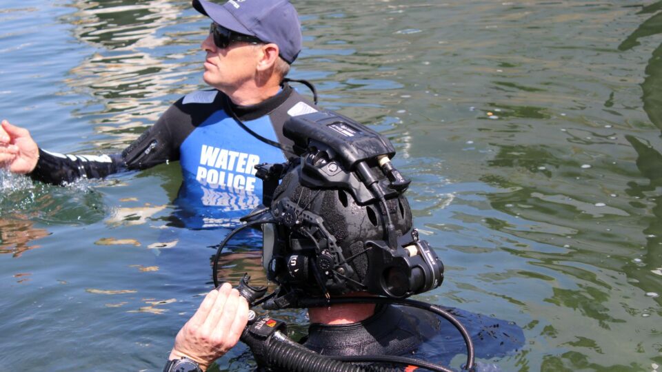 Two divers in open water: one wearing a blue 'Water Police' vest and cap, the other in a heavy diving helmet and breathing gear.