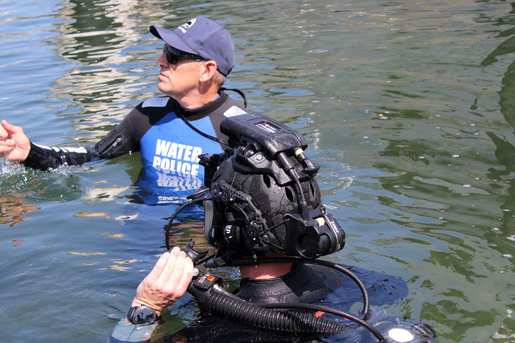 Two divers in open water: one wearing a blue 'Water Police' vest and cap, the other in a heavy diving helmet and breathing gear.